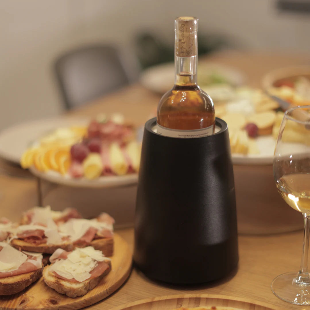 A wine bottle in a sleek black wine chiller on a wooden dining table, surrounded by appetizers, fruit platters, and a glass of white wine.