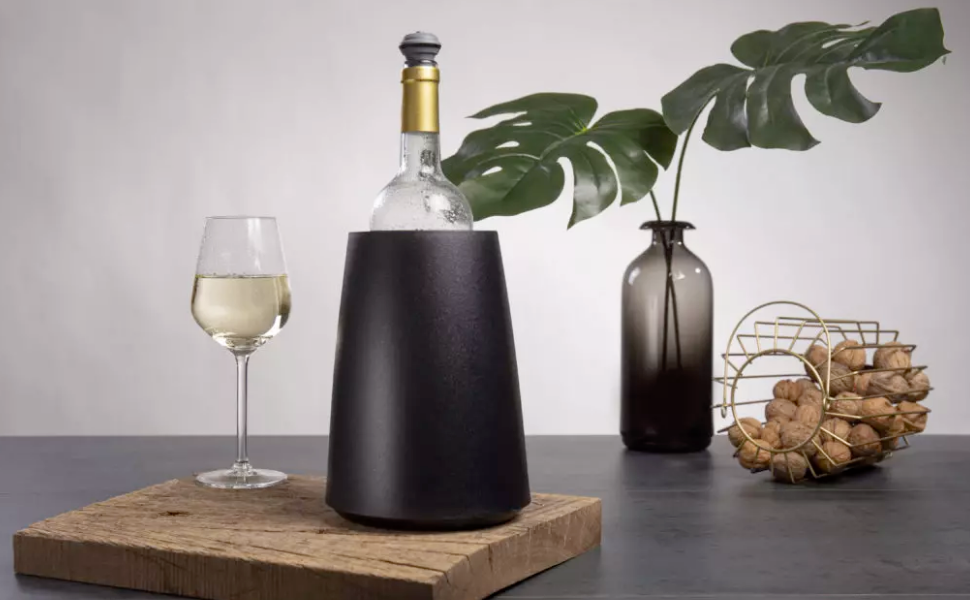 A chilled white wine bottle in a sleek black wine cooler, next to a glass of white wine on a wooden board, with decorative elements including a vase with green leaves and a wire basket of walnuts in the background.