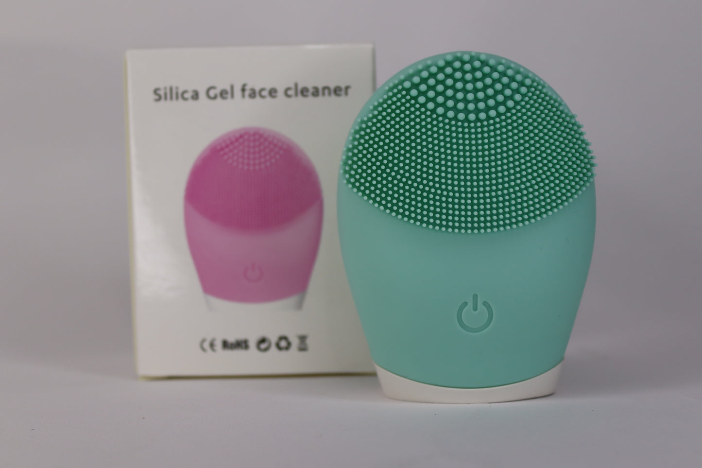 A green silicone facial cleansing brush with soft bristles and a power button, placed next to its packaging box labeled "Silica Gel Face Cleaner".