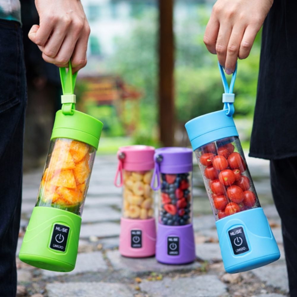 Two people holding portable 380ml mini blenders (green and blue) filled with fruit, with pink and purple blenders in the background outdoors.